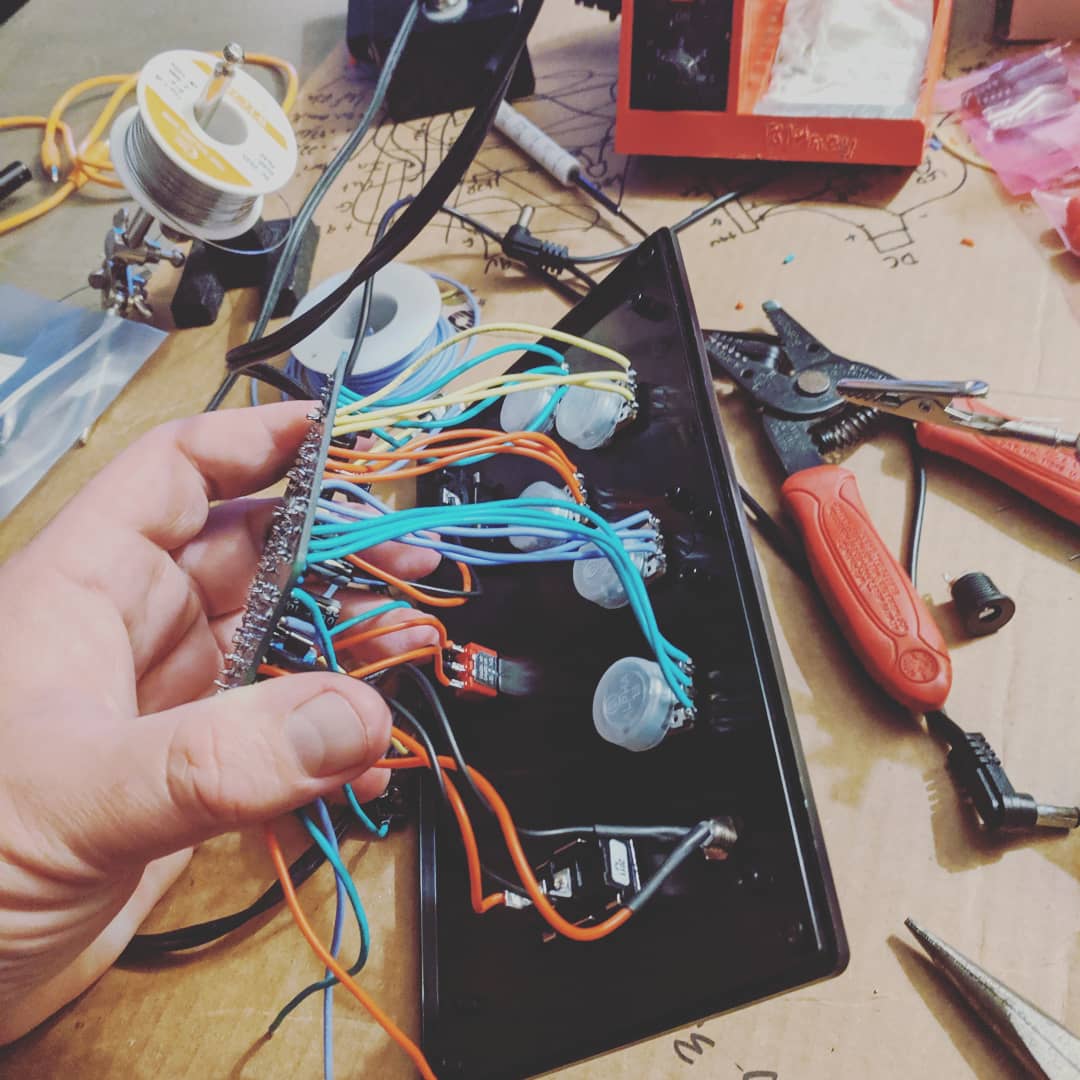 A hand holds a black plastic plate with multi-colored wires attached to various electronic components. In the background there is equipment for soldering and a wire cutter.